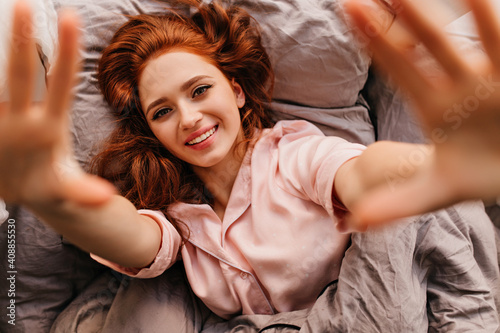 Merry ginger girl lying in bed with smile. Overhead shot of pleasant young woman posing in morning.