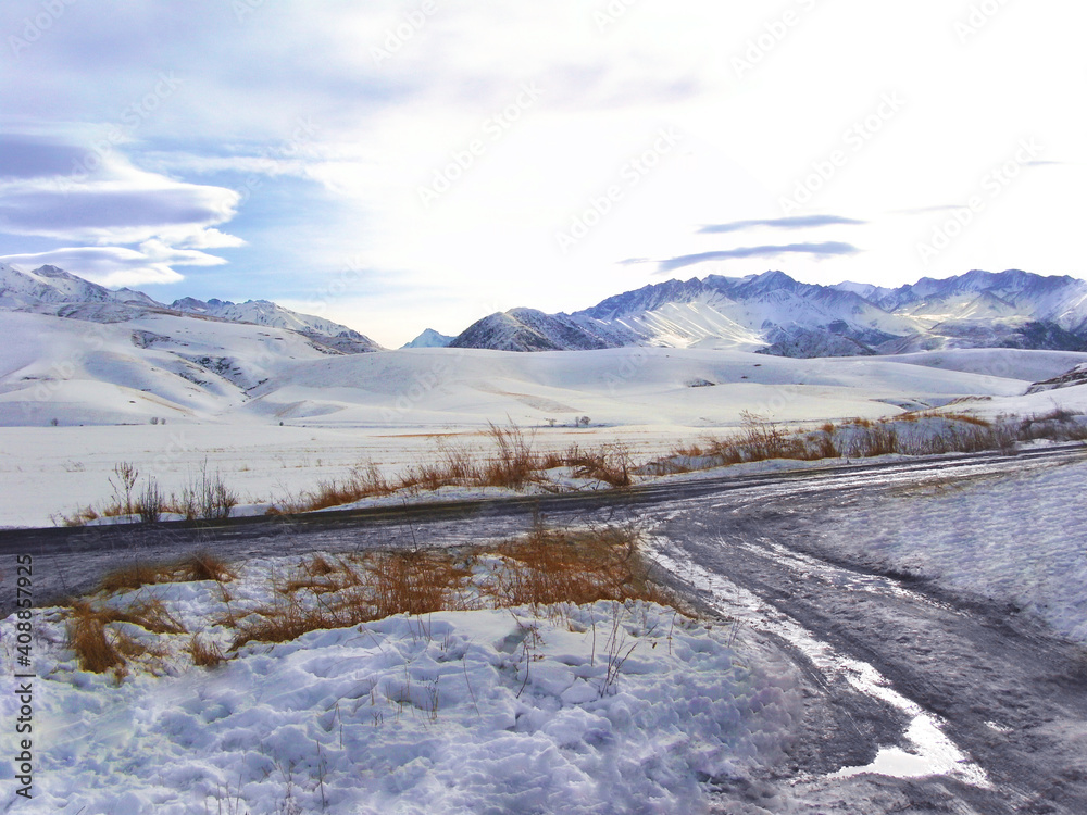 Snowy mountains and ridge, country road with puddle, brown grass in the foreground. Winter landscape. Copy space