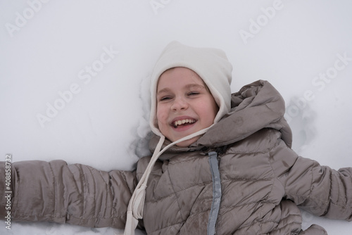 The child lies in deep snow and smiles sweetly. A beautiful girl in a snow-white environment. Concept for winter holidays and walks. Place for text
