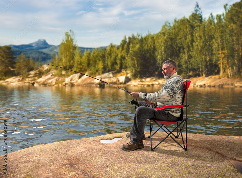 A Norwegian sits in a chair by a lake on a rock. He's got a fishing rod in his hands. In the background are waves and the landscape with mountains. A sunny afternoon.