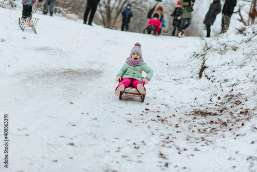A small and beautiful girl, a child, dressed in warm clothes, in the cold winter goes down the hill on a wooden, metal sled through the fresh white snow. Emotional photography.