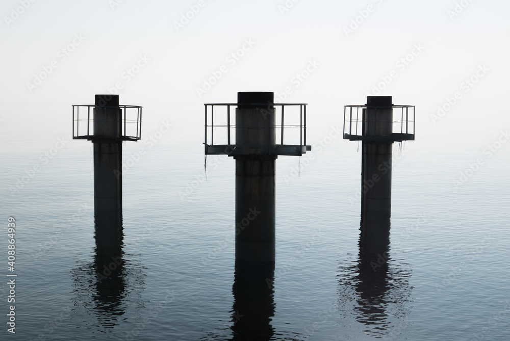 Three huge pillars of concrete emerge from the water against a strong diffused light, with reflections on water surface.