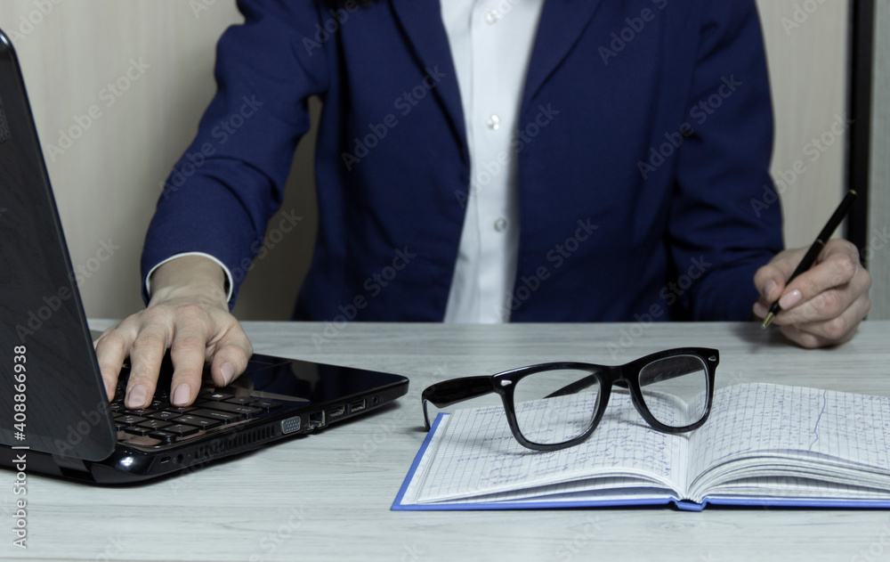 Side view. Young business woman sitting at table and taking notes in notebook.On table is laptop and glasses . Side view. Young business woman sitting at table and taking notes in notebook.On table is laptop and glasses .