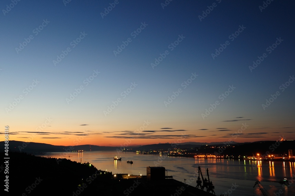 View from a height to Murmansk and the port against the background of mountains, sunset, sailing ships and their reflection in the water. Sea sunset in the port of Murmansk, Russia