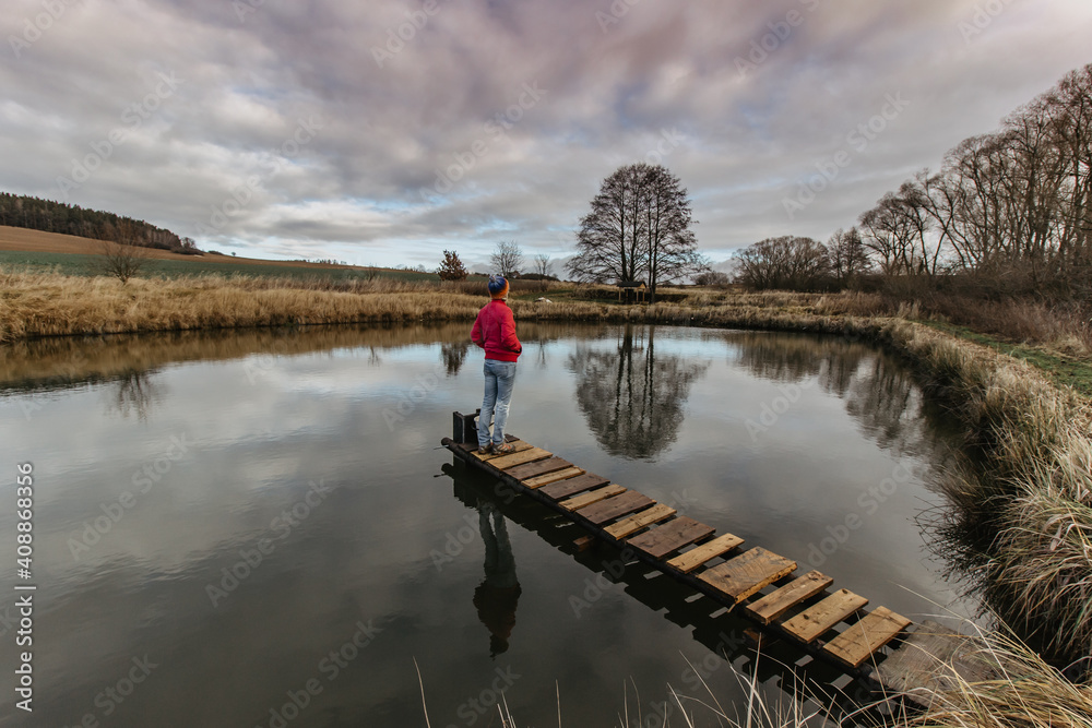 Woman watching reflections in pond during cold winter morning in the countryside.Girl in a pink jacket enjoying winter afternoon in the nature. Water and clouds. Wooden pier on lake. Tranquil scene