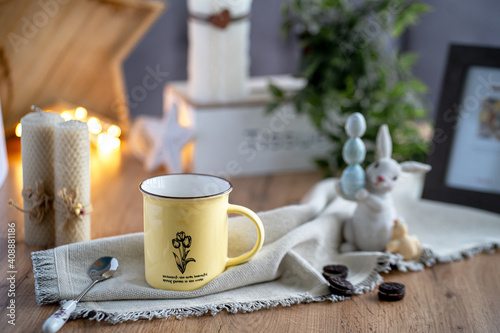 on the table there is a tea composition consisting of a yellow tin cup  a teaspoon  rows of Easter hare with Easter eggs  in the background a picture and candles. 