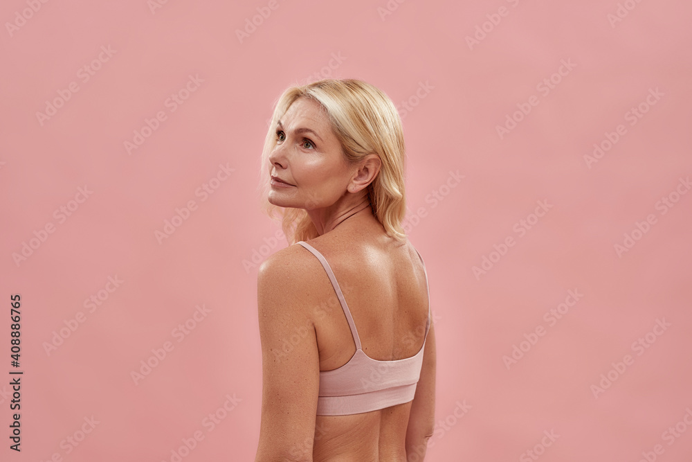Aging process. Rear view of a beautiful middle aged caucasian blonde woman wearing underwear looking away while posing isolated over pink background Aging process. Rear view of a beautiful middle aged caucasian blonde woman wearing underwear looking away while posing isolated over pink background