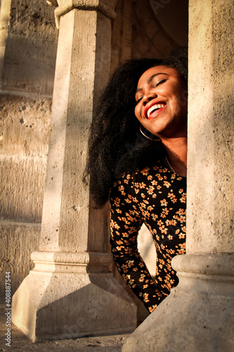 Black woman in a black dress with flowers and red lips smilling joyfully at the light of a sunrise between to columns made of stone