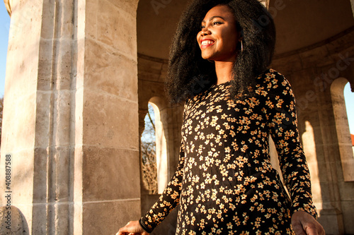 Black woman in a black dress with flowers and red lips smilling joyfully at the light of a sunrise