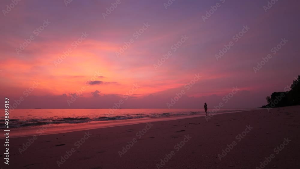 Lonely beach after sunset, bright red colored afterglow on sky. Tourist woman wearing swimsuit, go along water, step in soft waves, relaxing after hot day. Tropical vacation concept Lonely beach after sunset, bright red colored afterglow on sky. Tourist woman wearing swimsuit, go along water, step in soft waves, relaxing after hot day. Tropical vacation concept