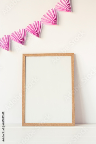 Empty wooden frame stands near white wall with pink paper garland hearts. Mockup poster frame close up in home interior decorated for Valentines day. Vertical image.