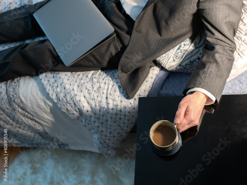 Coffee break working from home man in a suit closed laptop sits on bed hand on coffee cup