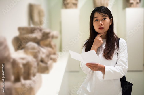 Young chinese woman visitor with guide book looking at exhibition in museum of ancient sculpture