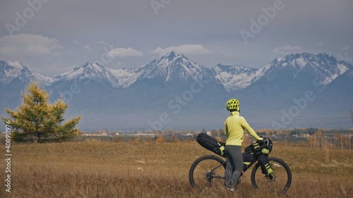 The woman travel on mixed terrain cycle touring with bikepacking. The traveler journey with bicycle bags. Sport tourism bikepacking, bike, sportswear in green black colors. Mountain snow capped.