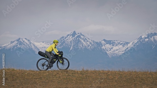 The woman travel on mixed terrain cycle touring with bikepacking. The traveler journey with bicycle bags. Sport tourism bikepacking, bike, sportswear in green black colors. Mountain snow capped.