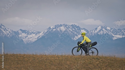 The woman travel on mixed terrain cycle touring with bikepacking. The traveler journey with bicycle bags. Sport tourism bikepacking, bike, sportswear in green black colors. Mountain snow capped.