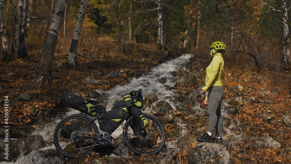 Obraz premium The woman travel on mixed terrain cycle touring with bikepacking. The traveler journey with bicycle bags. Sportswear in green black colors. The trip in magical autumn forest, river stream.