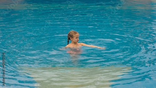 The little cute girl have fun in the swim pool. The child enjoy summer vacation in a swimming pool jumping, spinning, splash water. Slow motion. Happy childhood.