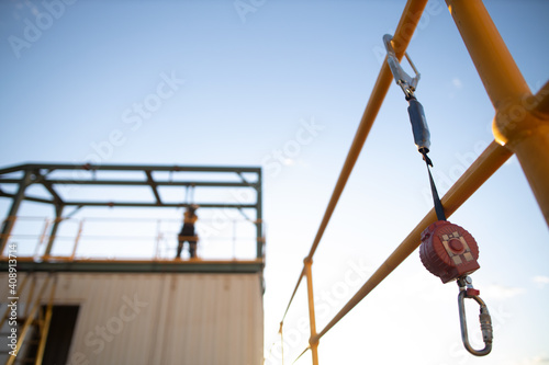 Safe work practices an inertia reel shock absorbing fall protection hook lanyard device clipping hanging on handrail with defocused construction worker working at heights in fall restraint background
