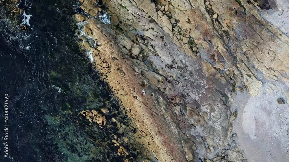 Slow aerial drone pan overhead of lone person standing on rocky foreshore with ocean waves slowly rolling in over rocks to reveal path. Slow aerial drone pan overhead of lone person standing on rocky foreshore with ocean waves slowly rolling in over rocks to reveal path.