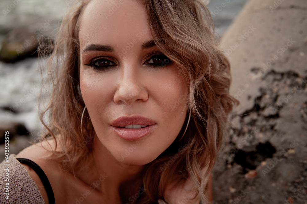 Portrait of beautiful sexy woman with smoky-eyes make-up sitting by the sea with dramatic stormy sky , enjoys the wind and look. Alone woman dressed black dress and beige cardigan Portrait of beautiful sexy woman with smoky-eyes make-up sitting by the sea with dramatic stormy sky , enjoys the wind and look. Alone woman dressed black dress and beige cardigan