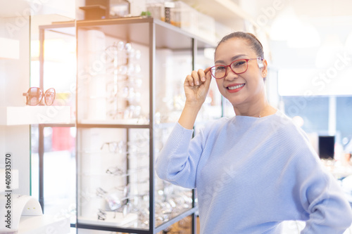 Middle aged asian woman choosing spectacles glasses in optician store. used correct or assist defective eyesight, smiling and looking at camera.