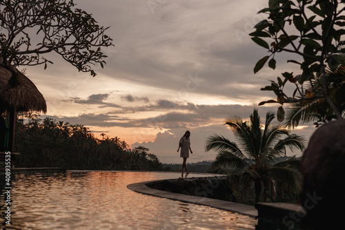 Silhoutte woman walking along the edge of a pool at sunset, coconut palm trees on background