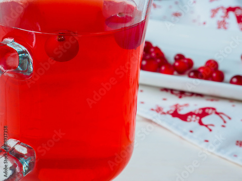 Selective focus on cranberries in a fresh drink in a glass cup. Berries on a white rectangular ceramic plate. Napkin with Christmas ornament. New Year's decor on a white wooden background. Copy space.