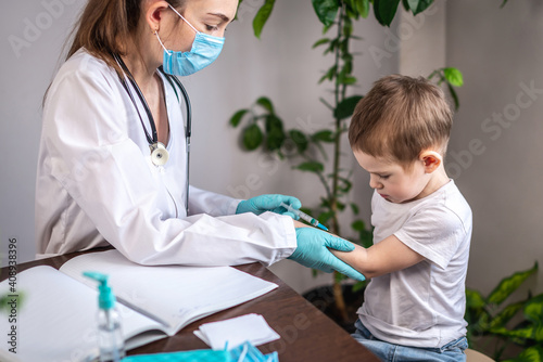 A young female doctor in a white medical coat  blue gloves  and a protective mask is vaccinating a small boy patient in his arm. Concept of health care and vaccination