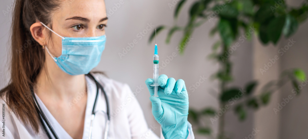 Young female doctor in a white gown, blue gloves, a protective mask and a stethoscope is holding a syringe with a vaccine in her hands. Concept of health care and vaccination Young female doctor in a white gown, blue gloves, a protective mask and a stethoscope is holding a syringe with a vaccine in her hands. Concept of health care and vaccination