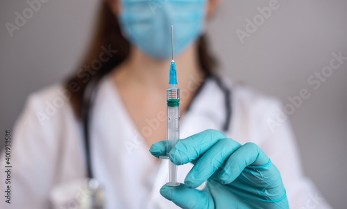 Young female doctor in a white gown, blue gloves, a protective mask and a stethoscope is holding a syringe with a vaccine in her hands. Concept of health care and vaccination. Closeup