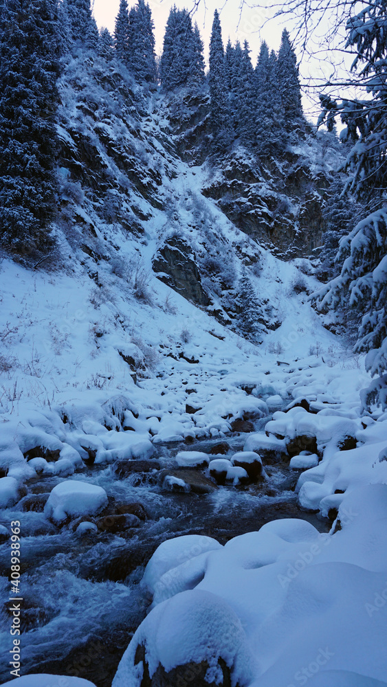 A clear mountain river runs through a snowy gorge. There is a metal bridge covered with snow. Tall spruce trees grow on the slopes of the mountains. There is steam from the river. Almarasan, Almaty A clear mountain river runs through a snowy gorge. There is a metal bridge covered with snow. Tall spruce trees grow on the slopes of the mountains. There is steam from the river. Almarasan, Almaty