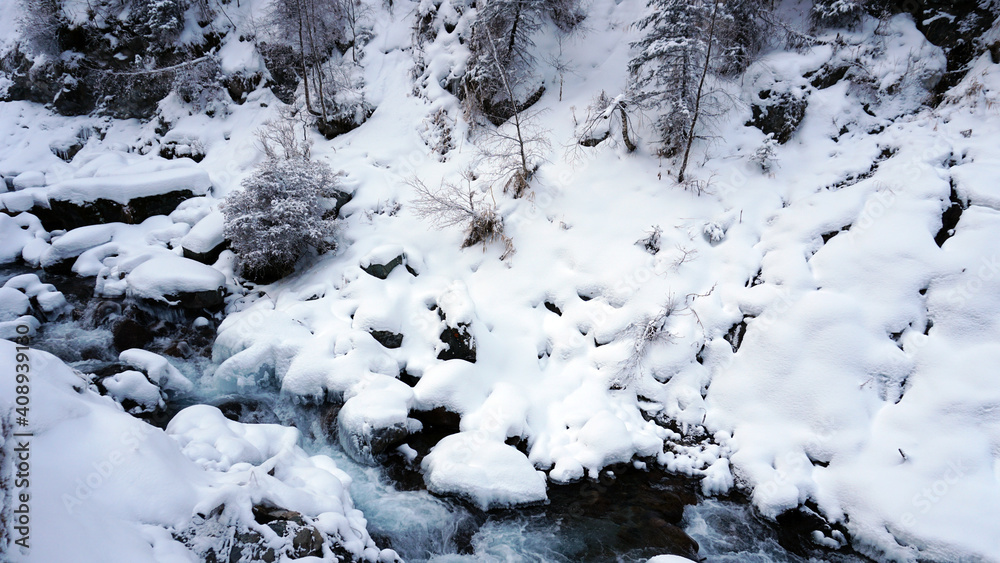 A clear mountain river runs through a snowy gorge. There is a metal bridge covered with snow. Tall spruce trees grow on the slopes of the mountains. There is steam from the river. Almarasan, Almaty A clear mountain river runs through a snowy gorge. There is a metal bridge covered with snow. Tall spruce trees grow on the slopes of the mountains. There is steam from the river. Almarasan, Almaty