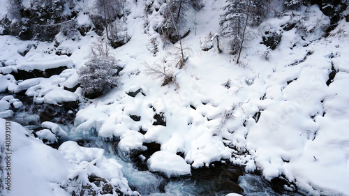 A clear mountain river runs through a snowy gorge. There is a metal bridge covered with snow. Tall spruce trees grow on the slopes of the mountains. There is steam from the river. Almarasan, Almaty © SergeyPanikhin