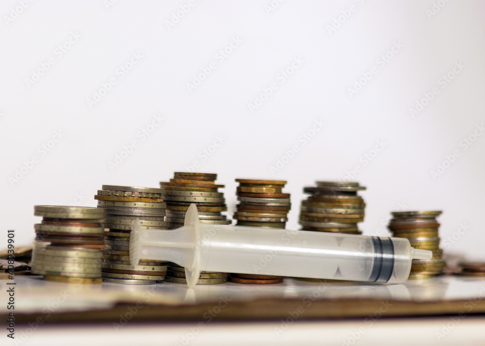 with metal coins of various shapes, countries and values, an empty medical syringe in the foreground with metal coins of various shapes, countries and values, an empty medical syringe in the foreground