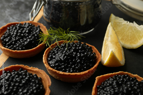 Board with tartlets and jar with caviar on black smokey background, close up