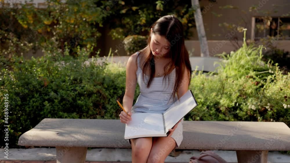Asian female college student working on her homework while setting on a bench outdoors Asian female college student working on her homework while setting on a bench outdoors