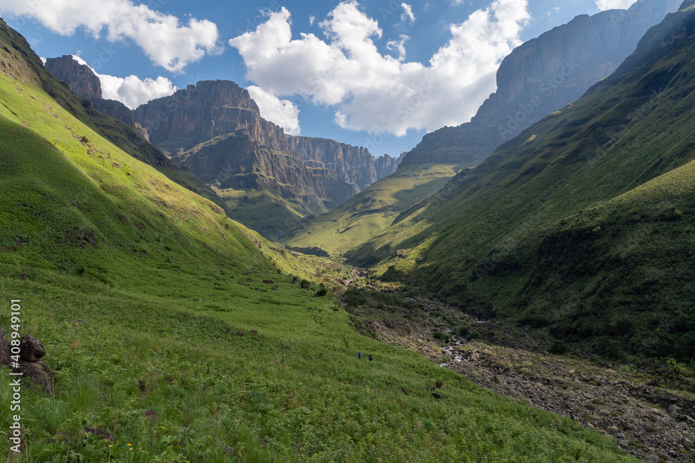 Hikers in lush valley below the towering escarment of the Drakensberg Mountains.