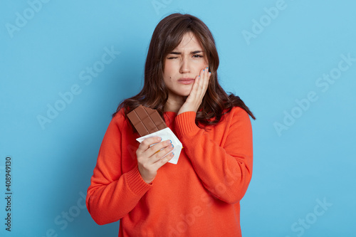 Woman suffering from toothache after eating chocolate, frowning her face, covering cheek with palm, female with dark hair wearing orange casual sweater.