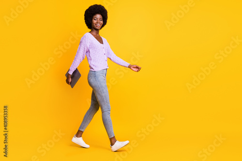 Full body profile photo of pretty dark skin person walking arm hold laptop isolated on yellow color background