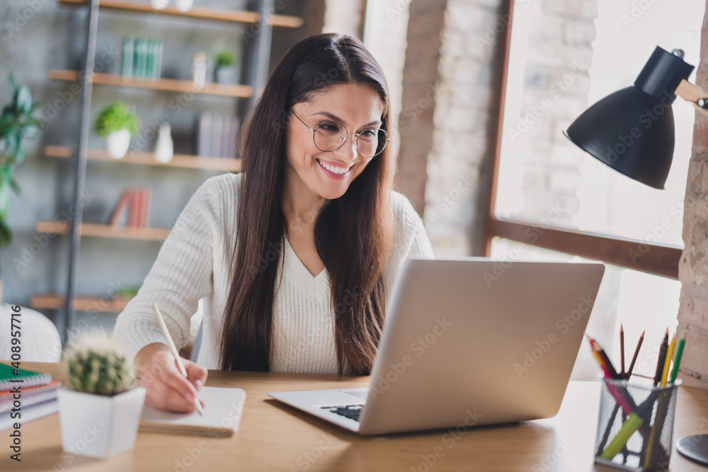 Photo of lady sit desk look screen laptop hold pen write notepad wear spectacles white sweater in living room home indoors Photo of lady sit desk look screen laptop hold pen write notepad wear spectacles white sweater in living room home indoors