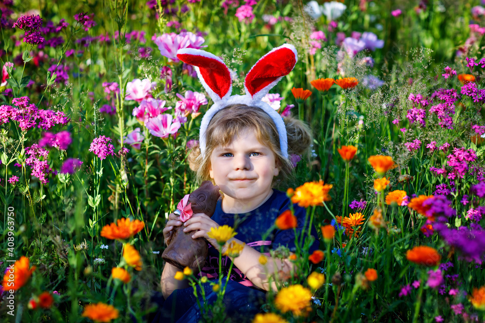 Portriat of adorable, charming toddler girl with Easter bunny ears eating chocolate bunny figure in flowers meadow. Smiling happy baby child on sunny day with colorful flowers, outdoors. Portriat of adorable, charming toddler girl with Easter bunny ears eating chocolate bunny figure in flowers meadow. Smiling happy baby child on sunny day with colorful flowers, outdoors.