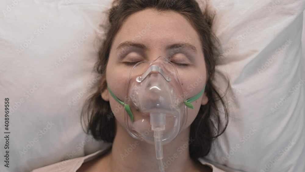 Close-up POV of young Caucasian brown-haired woman, lying head on pillow, wearing oxygen mask with eyes closed, then opening them and looking on camera