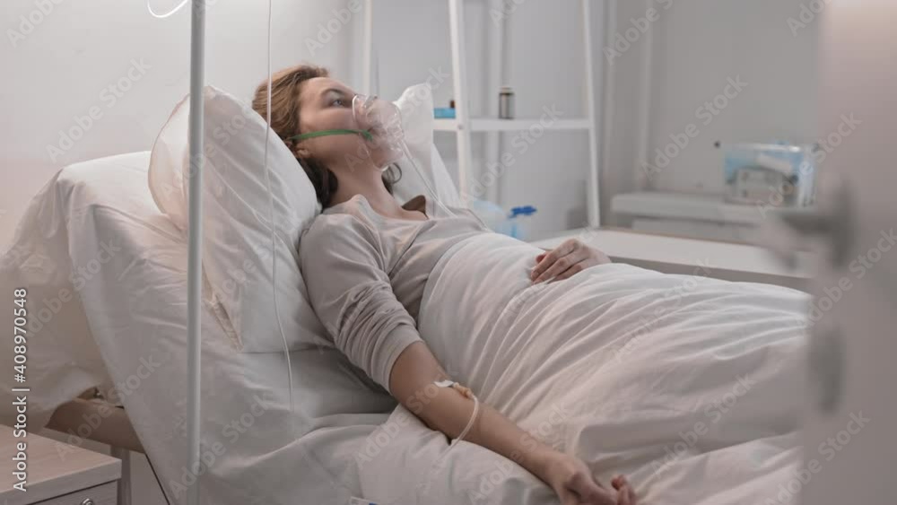 Side view medium long of young Caucasian brown-haired woman, lying in hospital bed, wearing oxygen mask and having dropper, then turning head and looking on camera Side view medium long of young Caucasian brown-haired woman, lying in hospital bed, wearing oxygen mask and having dropper, then turning head and looking on camera