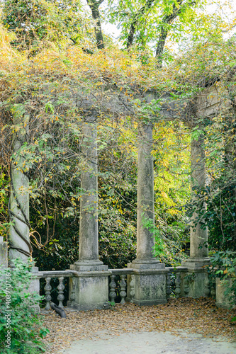 A neglected tropical garden with marble stone columns and a fence. The columns are overgrown with vines.