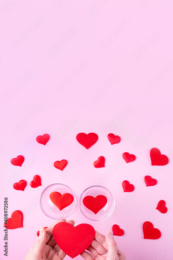 Hands hold a red heart shape, two glasses with hearts and white and red heart shapes on a pink background, top view, copy space, vertical frame. Valentine's Day concept