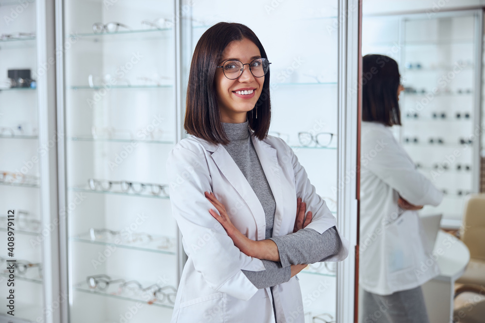 Good looking positive female in eyeglasses posing in room indoors in front of stand with tool for eye vision