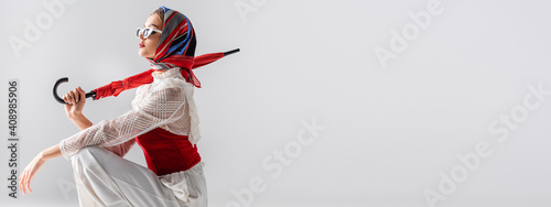 stylish woman in headscarf and sunglasses holding red umbrella while sitting isolated on white, banner