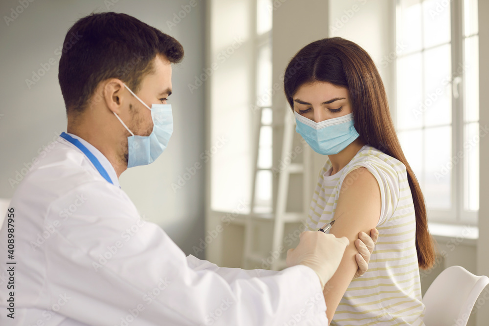 Fototapeta premium Male nurse or doctor in medical face mask giving flu injection to female patient during seasonal immunization campaign at health center. Young woman getting Covid-19 vaccine shot at clinic or hospital