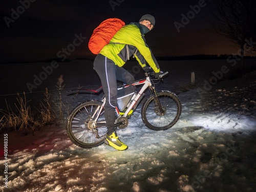 Mountain biker ride alone in snowy winter night. Tall biker wear green black thermal suit for winter cycling is ridding in snow.  Cold winter weather and snowy landscape. photo